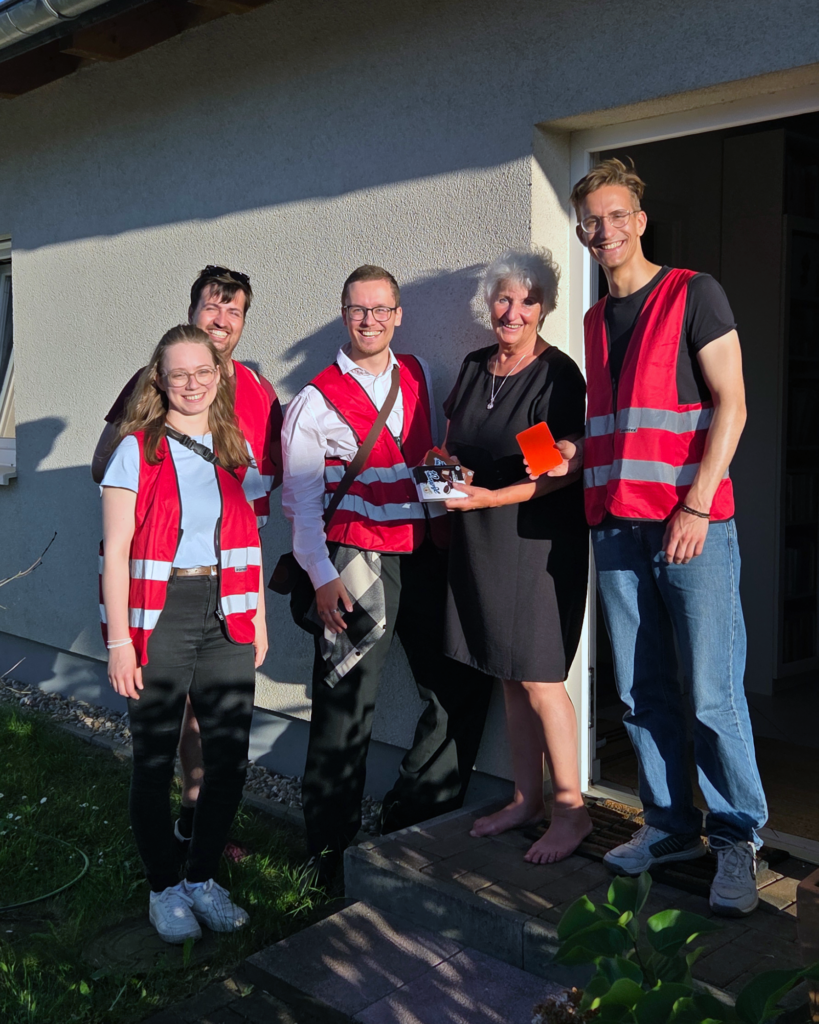 Vier Personen in Warnwesten stehen vor einer Haustür mit der Bewohnerin, die eine Packung Halorenkugeln in den Händen hält, die sie als Geschenk für die 5.000. Haustür erhalten hat.